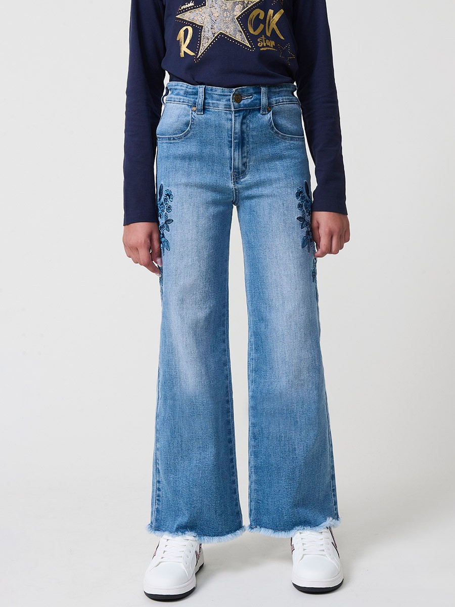 Pantalón de niña en denim elástico azul medio, pierna ancha, bajos deshilachados, cinco bolsillos y cintura regulable.