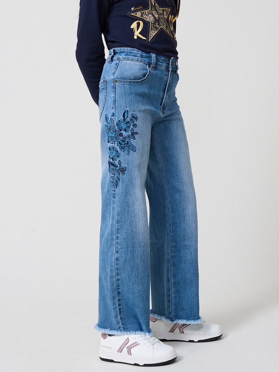 Pantalón de niña en denim elástico azul medio, pierna ancha, bajos deshilachados, cinco bolsillos y cintura regulable.