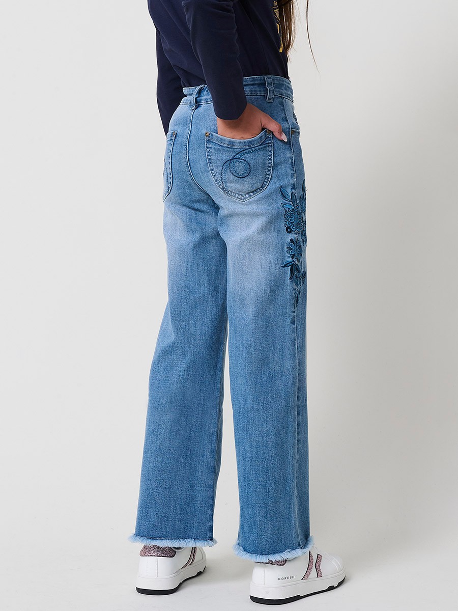 Pantalón de niña en denim elástico azul medio, pierna ancha, bajos deshilachados, cinco bolsillos y cintura regulable.