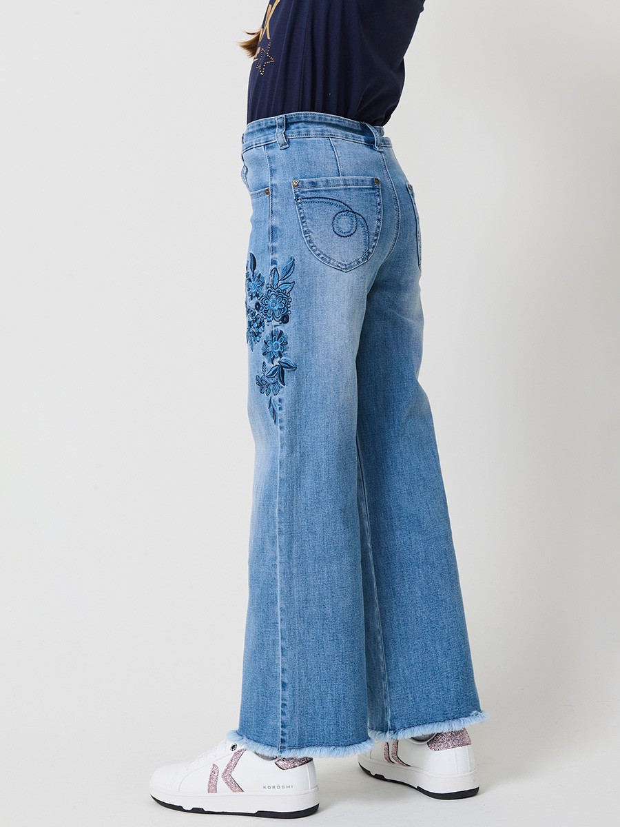 Pantalón de niña en denim elástico azul medio, pierna ancha, bajos deshilachados, cinco bolsillos y cintura regulable.