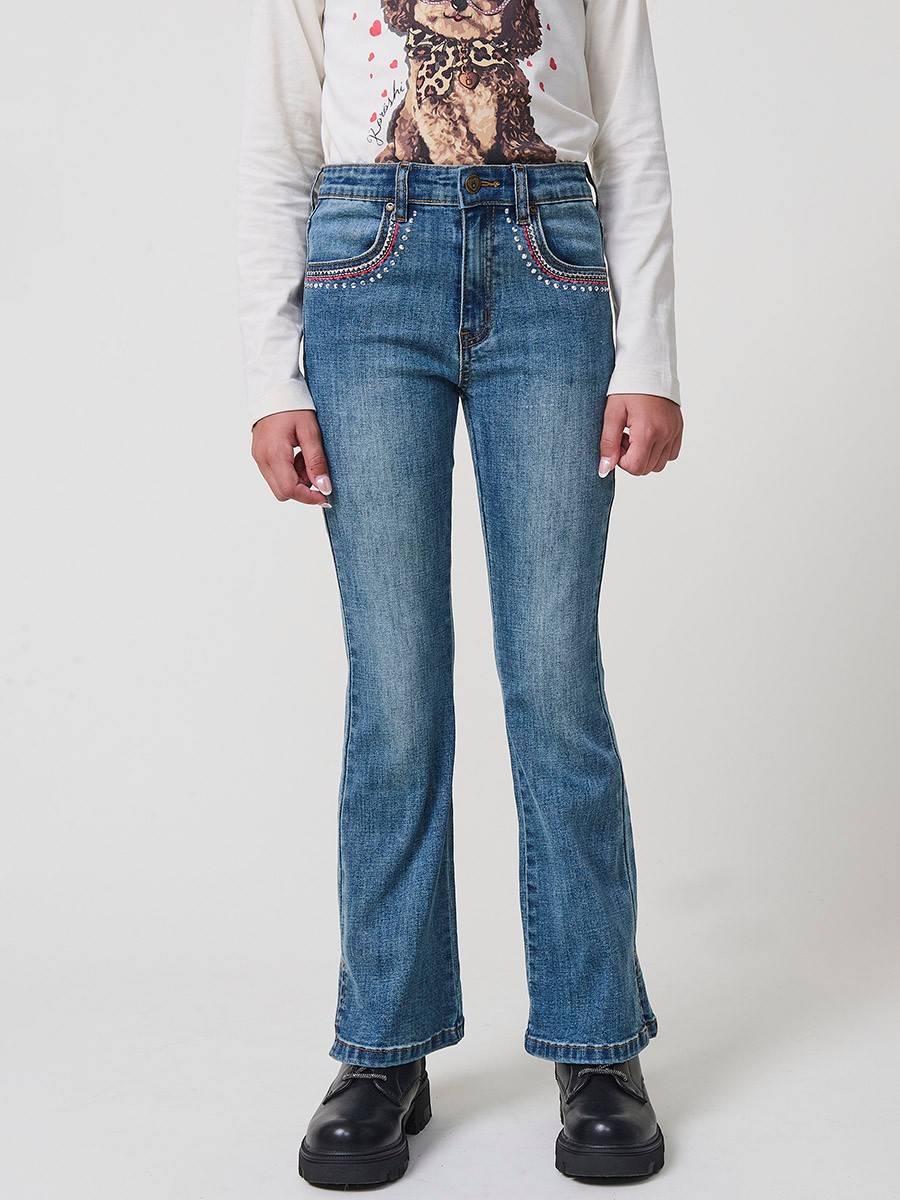 Pantalón de niña en denim elástico azul medio, pierna acampanada con cortes laterales, cinco bolsillos y cintura regulable.