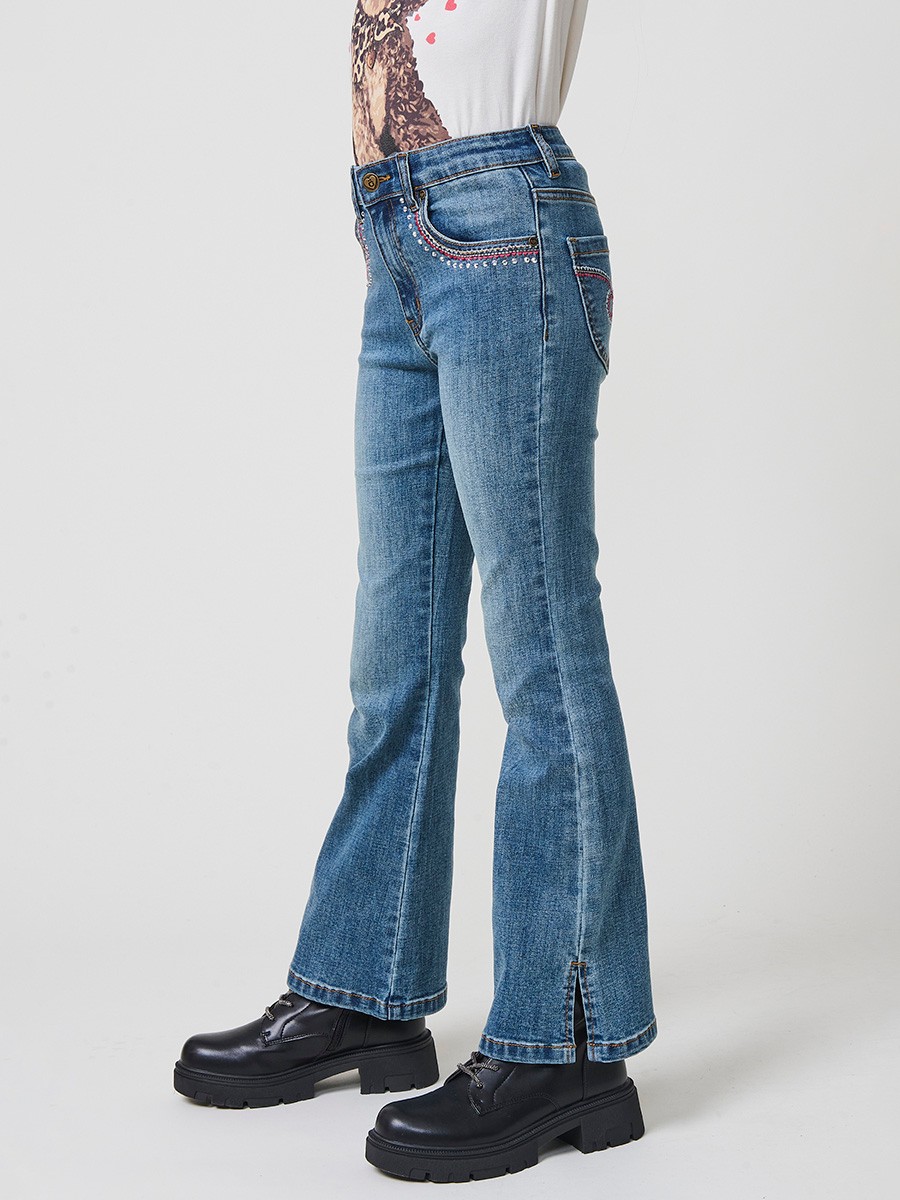 Pantalón de niña en denim elástico azul medio, pierna acampanada con cortes laterales, cinco bolsillos y cintura regulable.