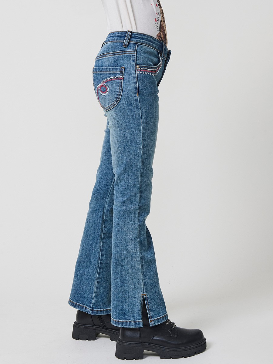 Pantalón de niña en denim elástico azul medio, pierna acampanada con cortes laterales, cinco bolsillos y cintura regulable.