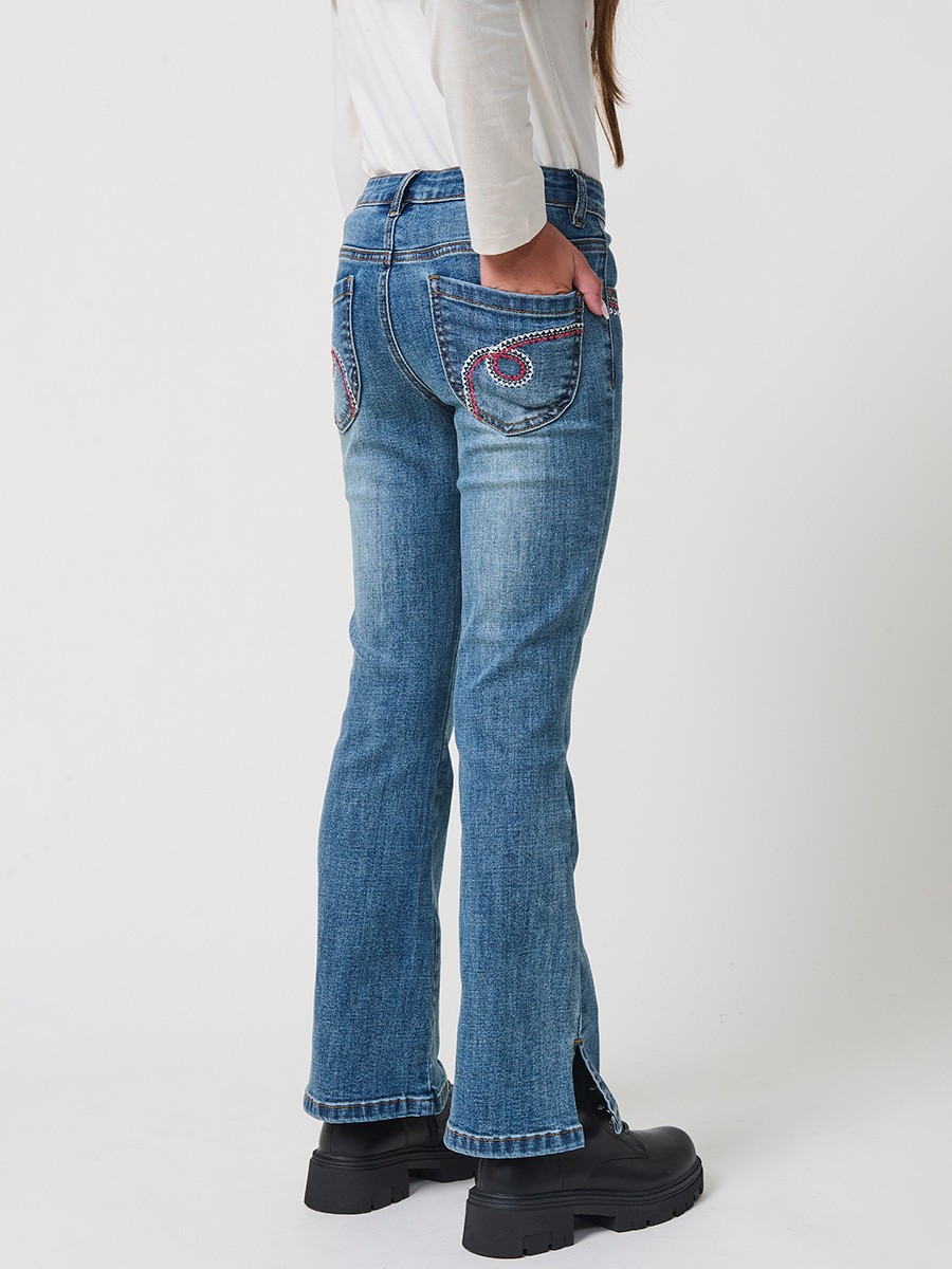 Pantalón de niña en denim elástico azul medio, pierna acampanada con cortes laterales, cinco bolsillos y cintura regulable.