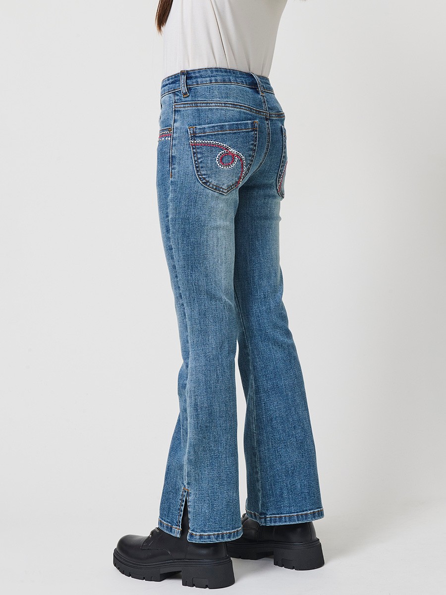 Pantalón de niña en denim elástico azul medio, pierna acampanada con cortes laterales, cinco bolsillos y cintura regulable.