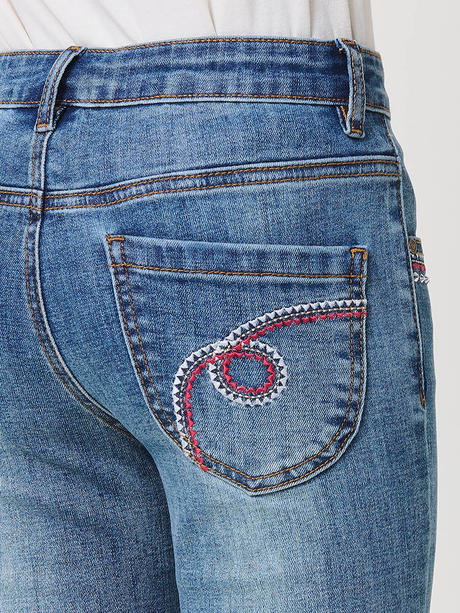 Pantalón de niña en denim elástico azul medio, pierna acampanada con cortes laterales, cinco bolsillos y cintura regulable.