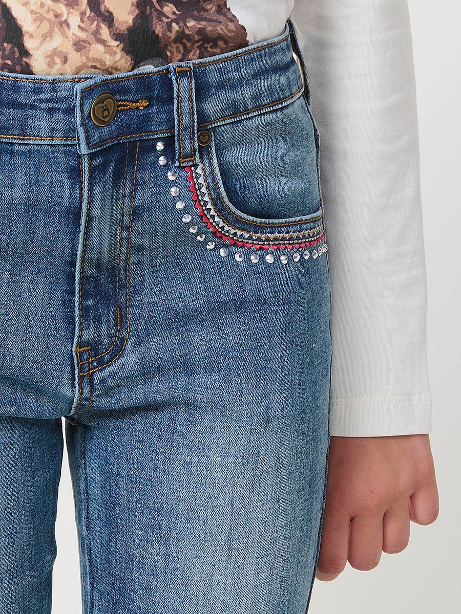 Pantalón de niña en denim elástico azul medio, pierna acampanada con cortes laterales, cinco bolsillos y cintura regulable.